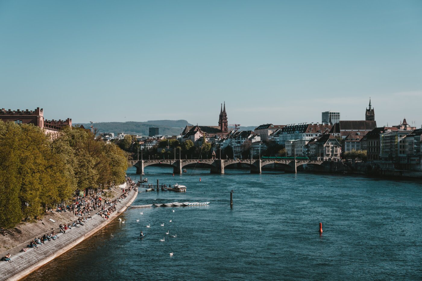 Ausblick über den Rhein. Links sonnen sich die Menschen am Flussufer, rechts sieht man die Basler Altstadt.