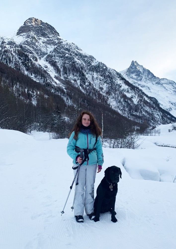 Josephine en combinaison de ski avec des bâtons de randonnée et son chien guide Quinto dans la neige.