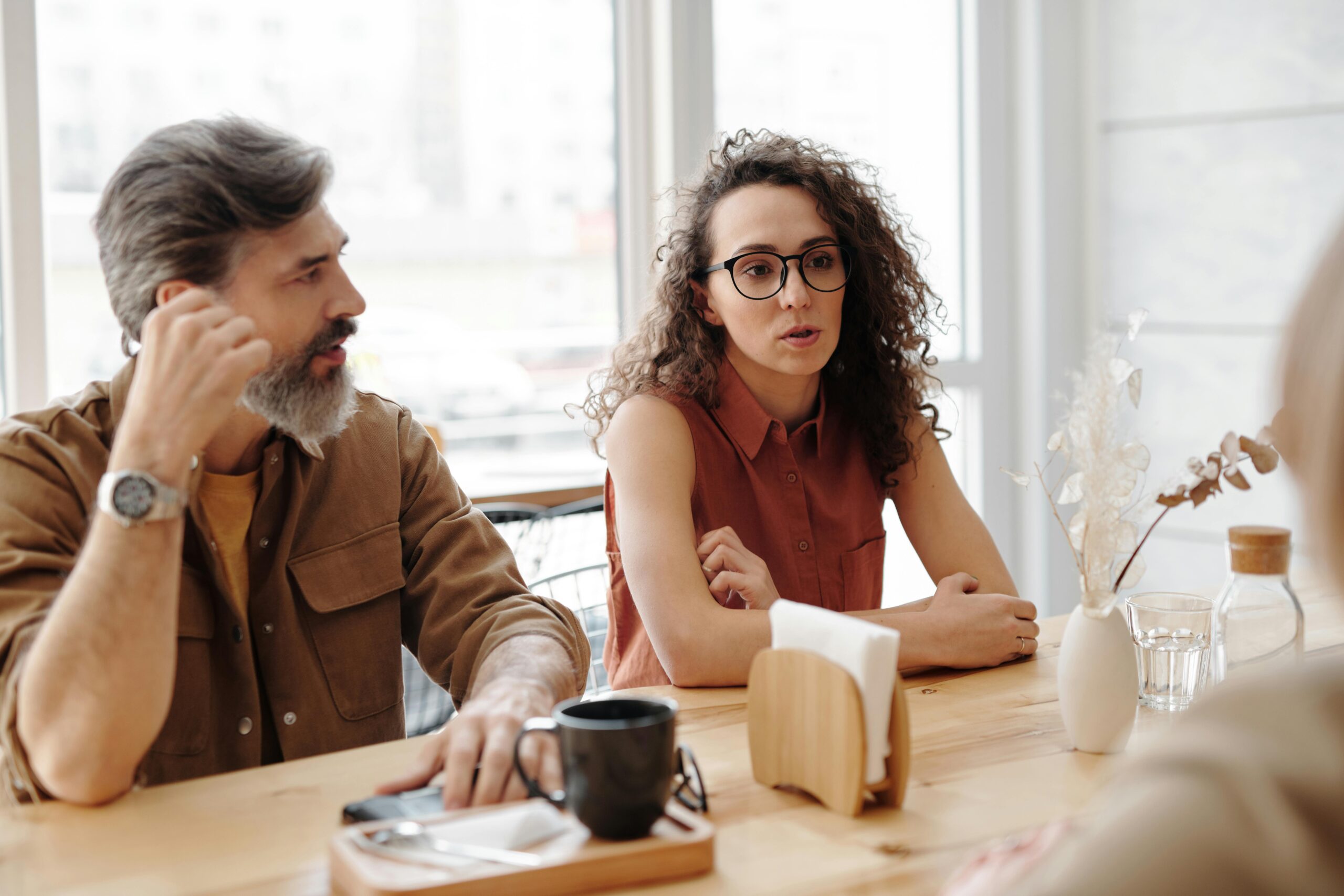 Un homme barbu et une femme aux cheveux bruns frisés en train de discuter avec d'autres personnes en dehors du tableau. Sur la table, il y a une tasse de café, un vase de fleurs et un porte-serviettes.