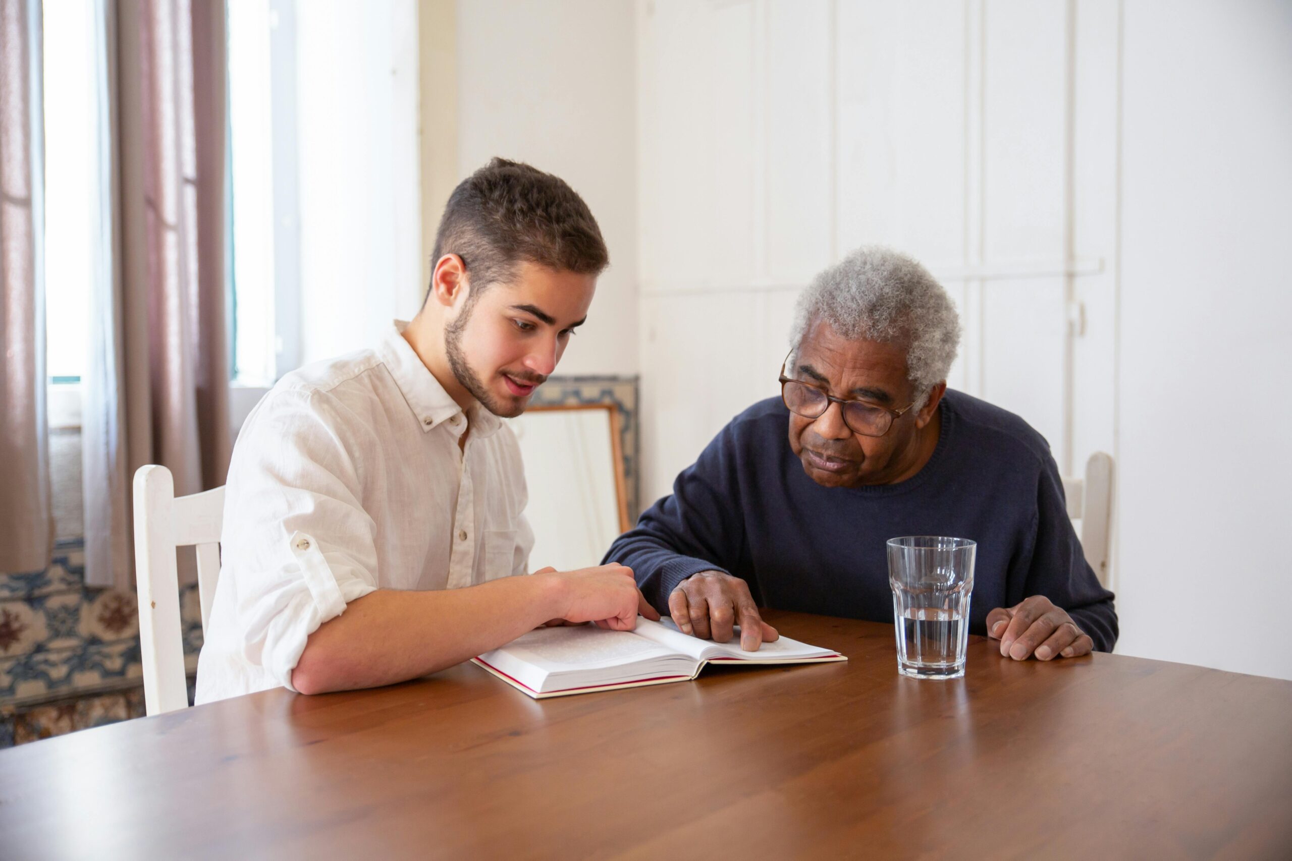 Un jeune homme aide une personne âgée à lire un livre à table.