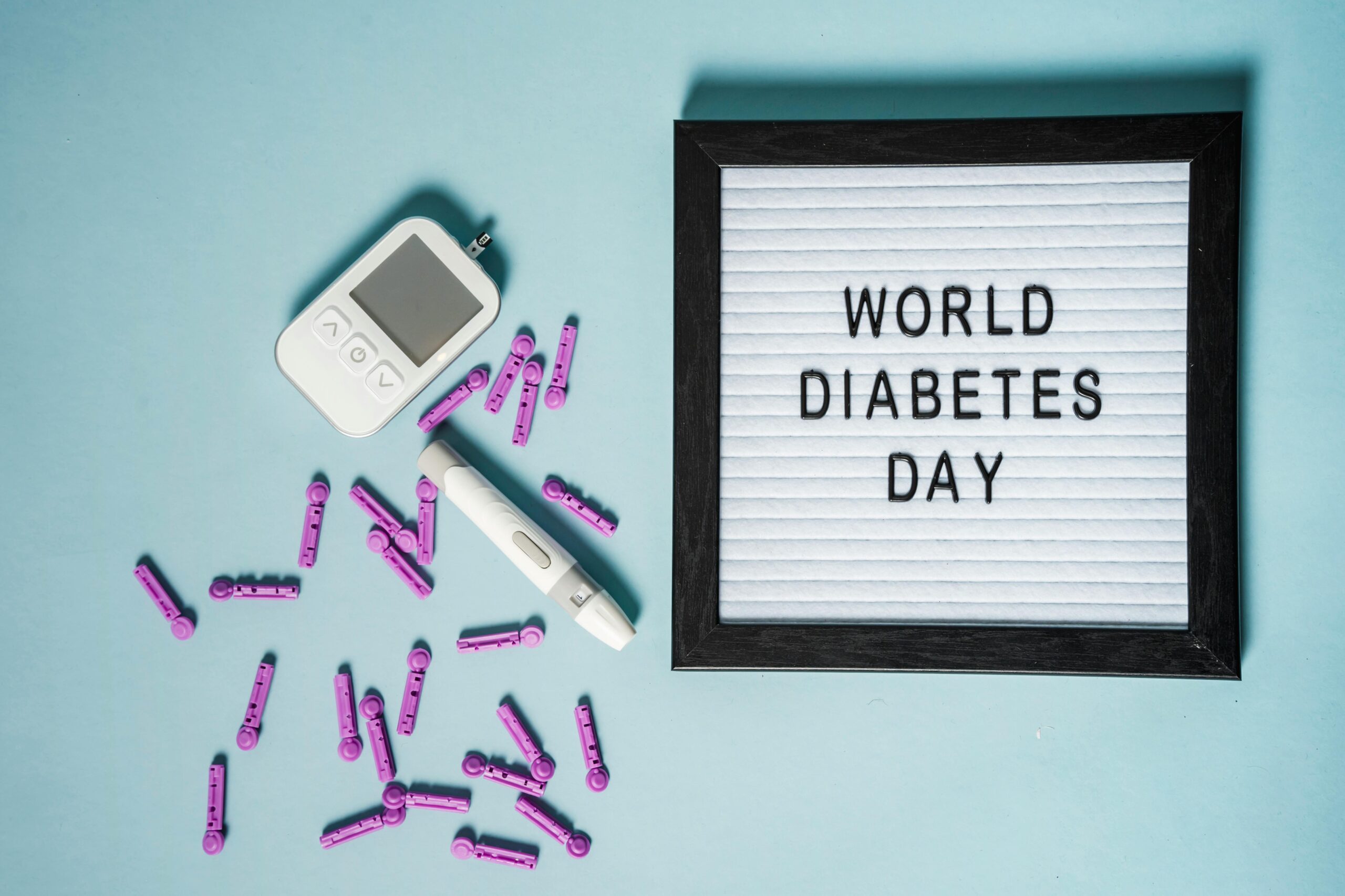 Auf einem blauen Hintergrund liegen ein Blutzuckermessgerät, eine Insulinspritze und viele Lanzetten sowie ein Bilderrahmen mit den Worten "World Diabetes Day" (Weltdiabetestag)