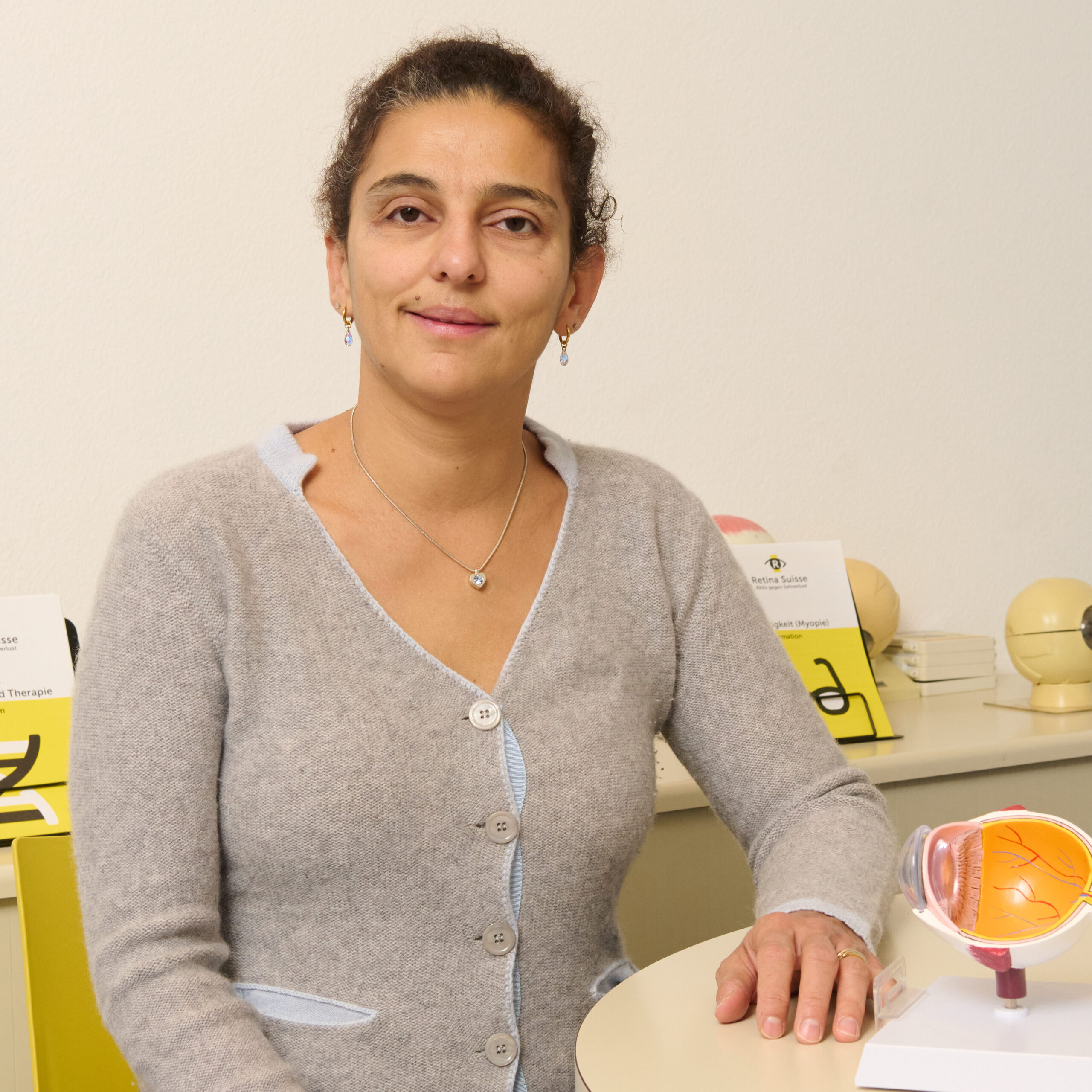 Rania Python est assise à une table ronde, le bras gauche posé nonchalamment sur la table. À côté d'elle se trouve un œil anatomique.