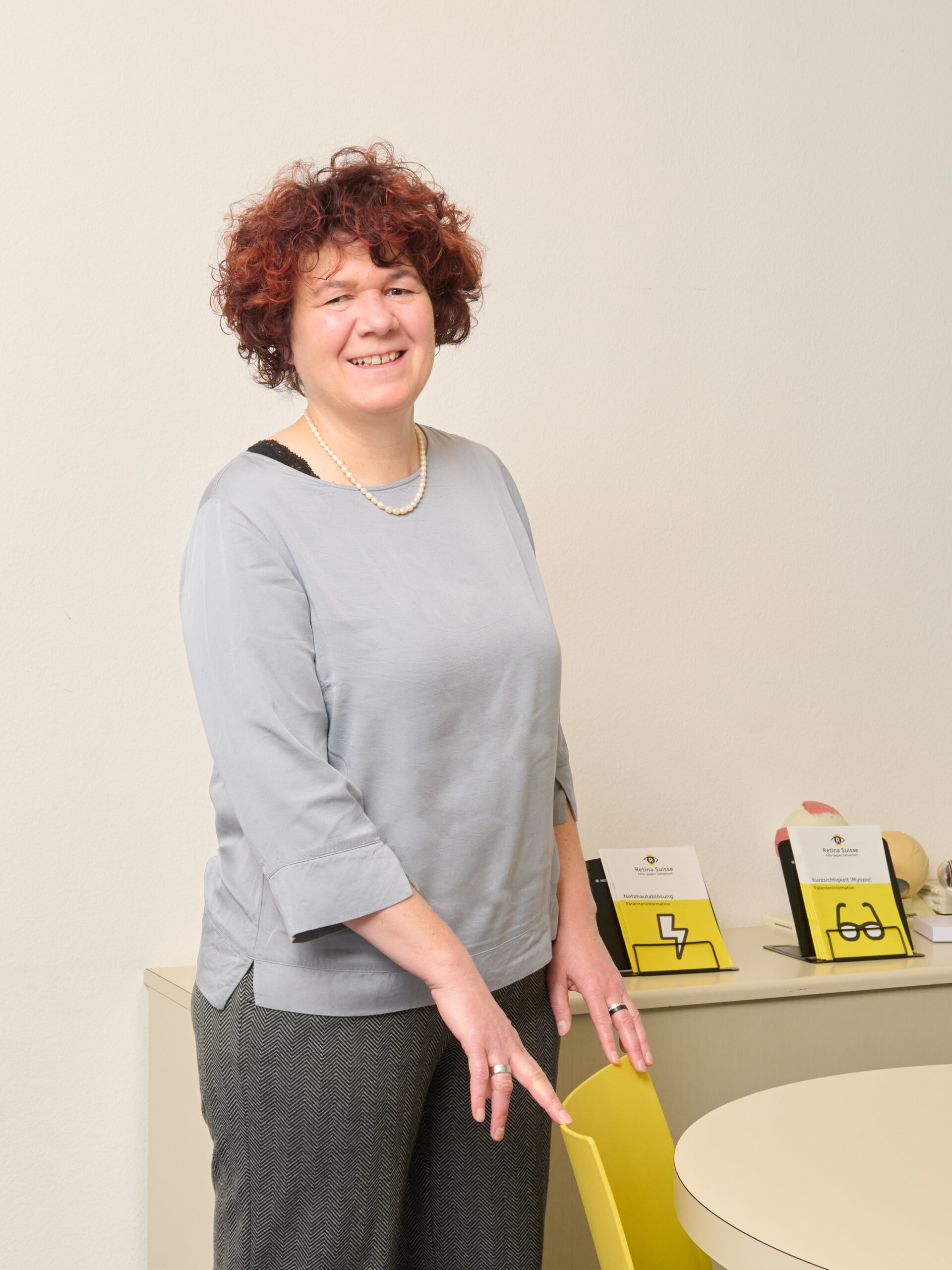 Susanne Trudel, Geschäftsleiterin von Retina Suisse, steht in ihrem Büro, die Hände ruhen locker auf der gelben Stuhllehne. Im Hintergrund sind Retina Suisse Informationsbroschüren zu sehen.