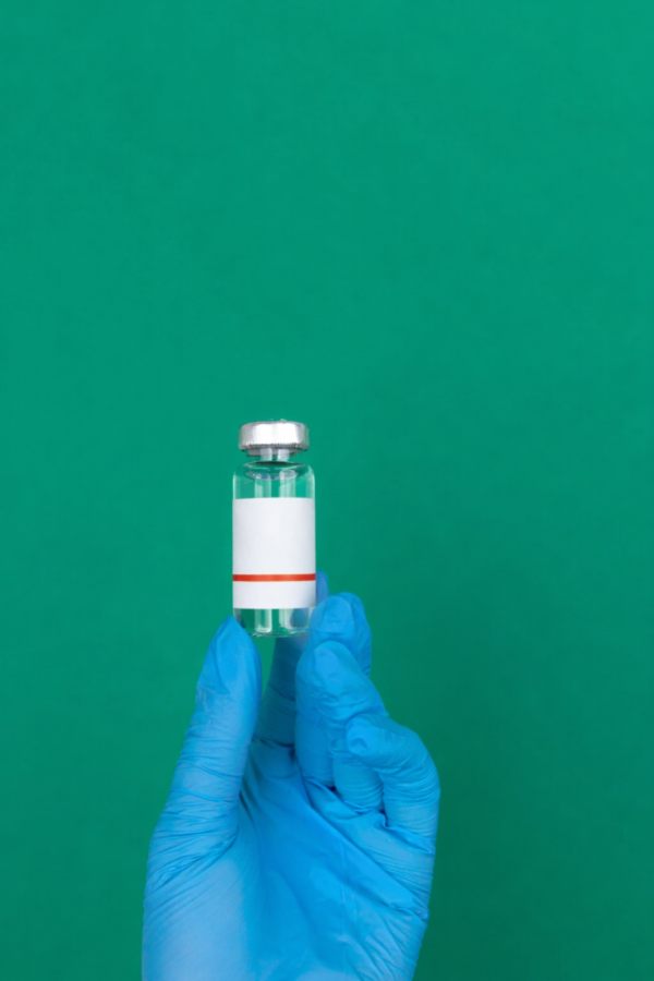 Bild: Eine blau behandschuhte Hand hält eine kleine durchsichtige Glasampulle mit silbernem Verschluss vor einem einfarbigen grünen Hintergrund. Die Ampulle enthält eine klare Flüssigkeit und hat ein weißes Etikett mit einem dünnen roten Streifen am unteren Rand.