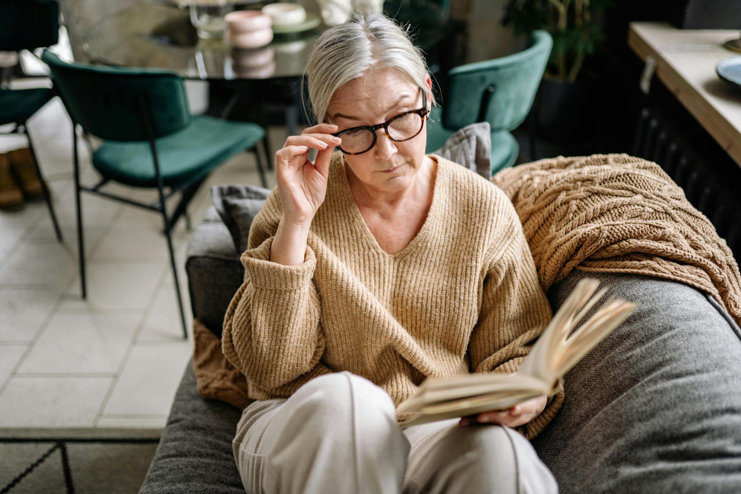 Eine Frau in ihren 50ern sitzt auf der Couch und liest ein Buch. Mit ihrer rechten Hand greift sie an die Seite ihrer Brille.